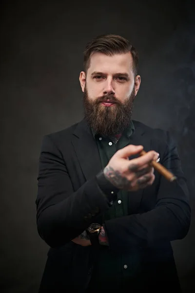 Brutal young businessman smokes a cigar in a dark studio. — Stock Photo, Image