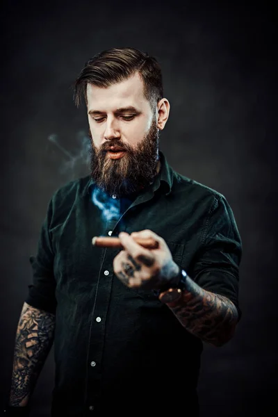 Studio portrait of a handsome tattooed hipster guy who smokes a cigar — Stock Photo, Image