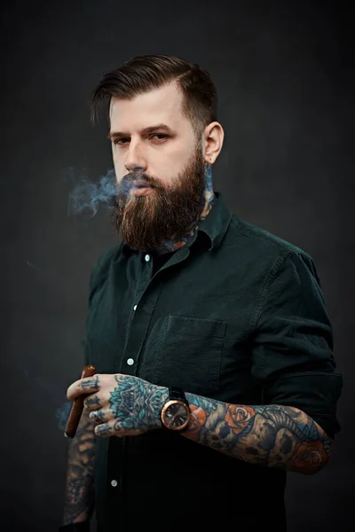 Studio portrait of a handsome tattooed hipster guy who smokes a cigar — Stock Photo, Image