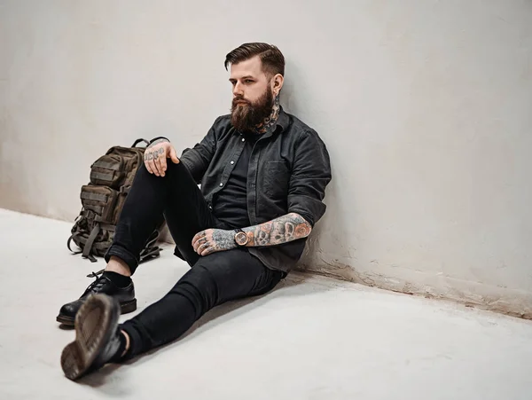 Bearded hipster guy with backpack sitting on a floor in studio — Stock Photo, Image