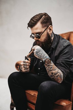 Havalı dövmeli sakallı adam eski bir sandalyede otururken puro yakıyor.