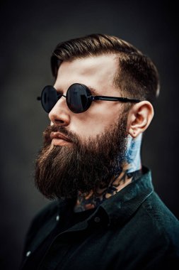 Koyu bir arka planda güneş gözlüklü havalı sakallı bir erkek portresi.