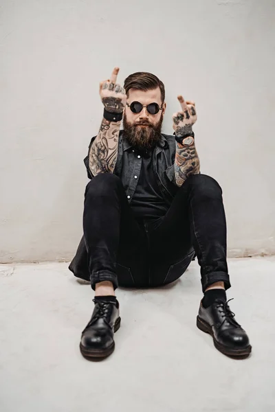Жорстокий бородатий хлопець, що показує середній палець робить хуй вираження, сидячи на підлозі — стокове фото