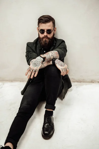 Cool bearded hipster guy in sunglasses sitting of a floor in studio — Stock Photo, Image