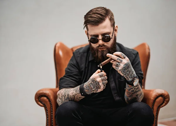 Cool tattooed bearded man lights a cigar while sitting on a vintage chair