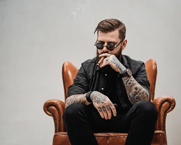 Τατουάζ γενειοφόρος άνδρας με κομψό κούρεμα στα γυαλιά ηλίου που καπνίζει ένα πούρο ενώ κάθεται σε μια vintage καρέκλα στο στούντιο — Φωτογραφία Αρχείου