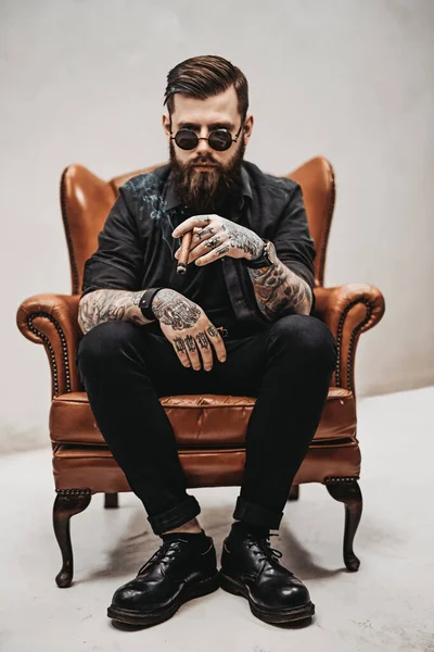 Homme barbu tatoué avec une coupe de cheveux élégante dans des lunettes de soleil qui fume un cigare tout en étant assis sur une chaise vintage en studio — Photo