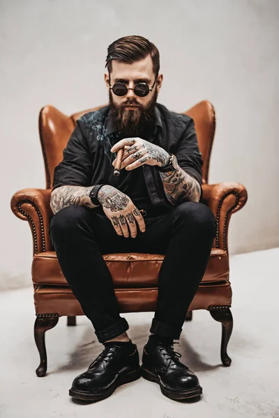 Tatuerad skäggig man med snygg frisyr i solglasögon som röker en cigarr sittande på en vintage stol i studio — Stockfoto