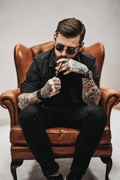 Homme barbu tatoué avec une coupe de cheveux élégante dans des lunettes de soleil qui fume un cigare tout en étant assis sur une chaise vintage en studio — Photo