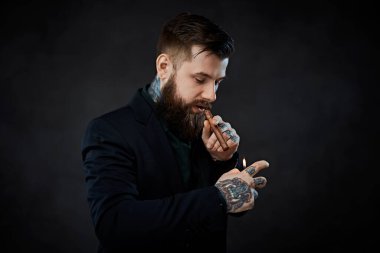 Zarif giyimli, sakallı, boynunda dövmeleri olan ve elinde puro yakan bir erkek.