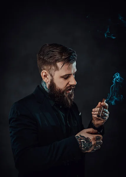Elegantemente vestido barbudo macho com tatuagens em seu pescoço e mão acende um charuto — Fotografia de Stock
