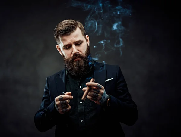 Brutal young businessman smokes a cigar in a dark studio. — Stock Photo, Image