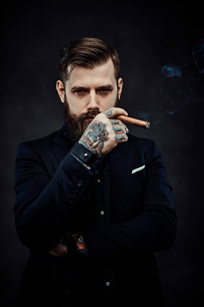 Жестокий молодой бизнесмен курит сигару в темной студии
.