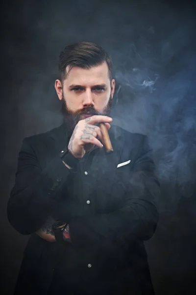 Жорстокий молодий бізнесмен тримає сигару і дивиться на камеру в димчастій темній студії — стокове фото