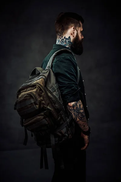 Vue arrière d'un hipster tatoué barbu avec sac à dos dans un studio sombre — Photo