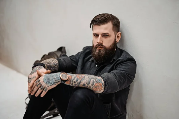 Bearded hipster guy with backpack sitting on a floor in studio — Stock Photo, Image