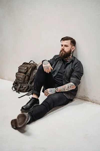 Bearded hipster guy with backpack sitting on a floor in studio — Stock Photo, Image