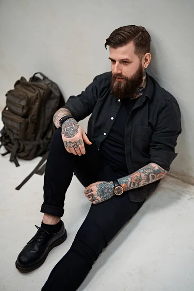 Бородатый хипстер с рюкзаком сидит на полу в студии — стоковое фото