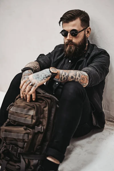 Бородатый хипстер в солнечных очках с рюкзаком, сидящим на полу в студии — стоковое фото