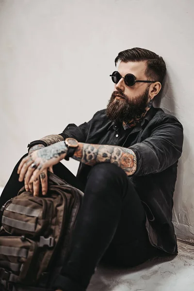 Brodaty hipster facet w okularach przeciwsłonecznych z plecakiem siedzi na podłodze w studio — Zdjęcie stockowe