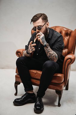 Havalı dövmeli sakallı adam eski bir sandalyede otururken puro yakıyor.
