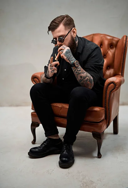 Cool tetované vousatý muž zapálí doutník, zatímco sedí na vinobraní židle — Stock fotografie