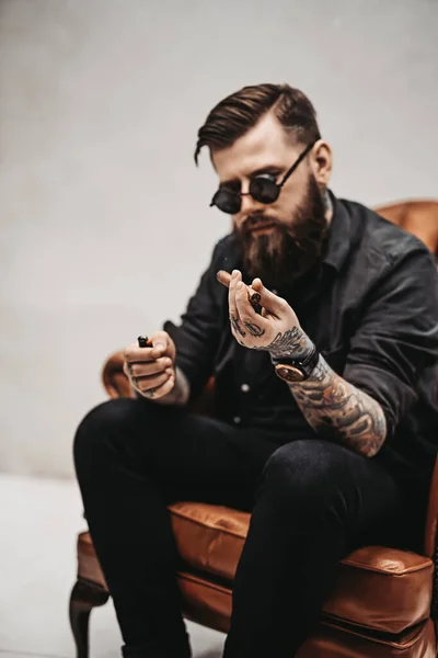 Cool tatuerad skäggig man tänder en cigarr medan du sitter på en vintage stol — Stockfoto