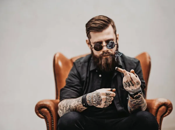 Homme barbu tatoué avec une coupe de cheveux élégante dans des lunettes de soleil qui fume un cigare tout en étant assis sur une chaise vintage en studio — Photo