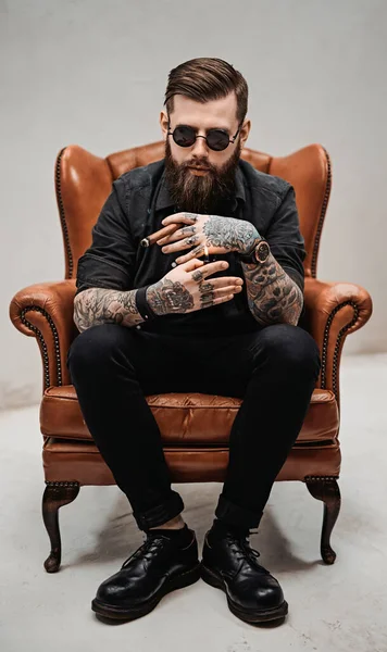 Tetovált szakállas férfi stílusos frizurával napszemüvegben, aki szivarozik, miközben egy vintage széken ül a stúdióban. — Stock Fotó
