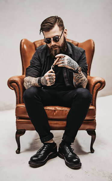 Tattooed bearded man with stylish haircut in sunglasses who smokes a cigar while sitting on a vintage chair in studio