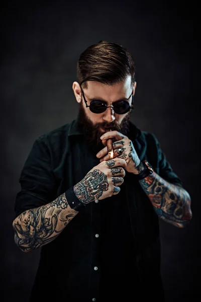 Bearded tattooed guy wearing green shirt and sunglasses lights a cigar — Stock Photo, Image