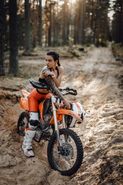 Motokros kıyafeti giyen, yarı çıplak gövdesiyle ormanda bisikletinin üstünde oturan bir kadın yarışçı.