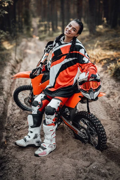 Bella donna corridore indossare protettivo motocross vestito sorridente e guardando sulla macchina fotografica mentre seduto sulla sua moto nel bosco — Foto Stock