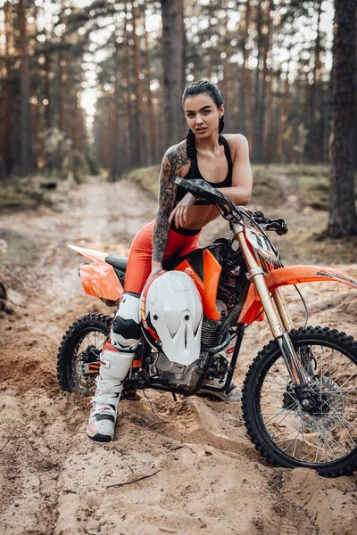 Femmina pilota indossa abito motocross con torso semi nudo seduto sulla sua moto nella foresta — Foto Stock