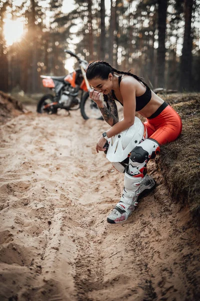 Brünettes Mädchen im Motocross-Outfit mit halbnacktem Oberkörper sitzt auf Gras und lacht hemmungslos — Stockfoto