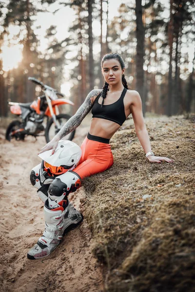 Krásná dívka nosí motokrosový outfit s polonahým trupem sedí na trávě v lese — Stock fotografie