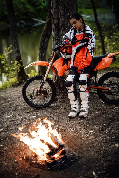 Motokros kıyafeti giymiş güzel bir bayan yarışçı ve ormanda bir şenlik ateşinin yanında ısınıyor. — Stok fotoğraf