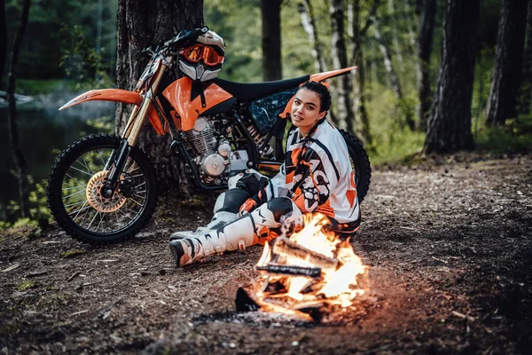 Urocza młoda zawodniczka w stroju motocrossa, rozgrzewająca się siedząc przy ognisku — Zdjęcie stockowe