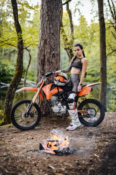 Motokros kıyafeti giymiş güzel bir bayan yarışçı yarı çıplak gövdesiyle ormanda şenlik ateşinin yanında oturuyor. — Stok fotoğraf