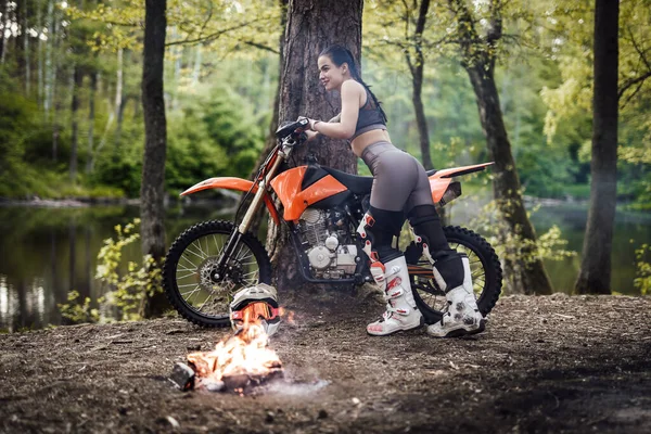 Urocza młoda zawodniczka w stroju motocrossa z półnagim tułowiem pochyla motocykl na drzewie obok ogniska w lesie — Zdjęcie stockowe