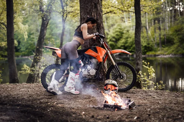 Encantadora joven corredor con traje de motocross con el torso semidesnudo inclina su motocicleta en un árbol junto a una hoguera en el bosque — Foto de Stock