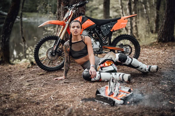 Charmig ung kvinnlig racer bär motocross outfit med halvnaken bål, värma upp medan sitter bredvid brasan — Stockfoto