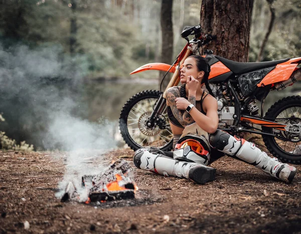 Urocza młoda zawodniczka w stroju motocrossa z półnagim tułowiem, rozgrzewająca się siedząc przy ognisku — Zdjęcie stockowe