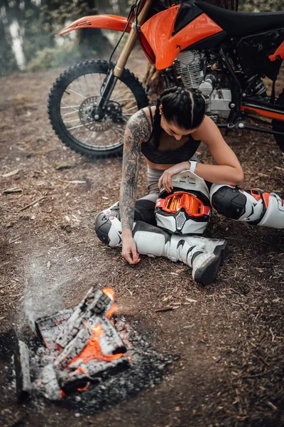 Motokros kıyafeti ve yarı çıplak gövde giyen çekici genç bayan yarışçı şenlik ateşinin yanında otururken ısınıyor. — Stok fotoğraf