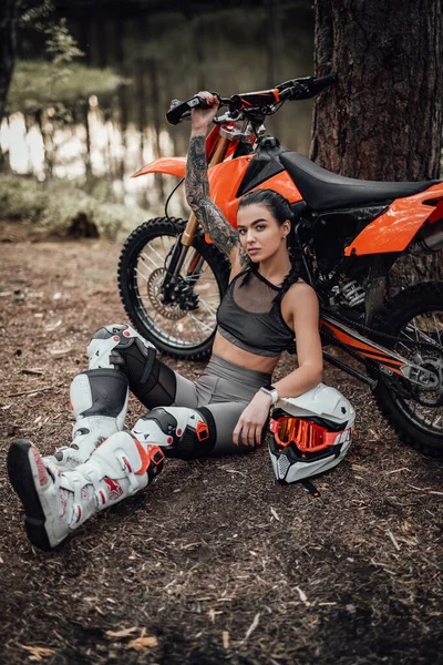 Sensuale ragazza corridore tatuato indossa abito motocross con torso semi nudo seduto accanto alla sua moto nel bosco — Foto Stock