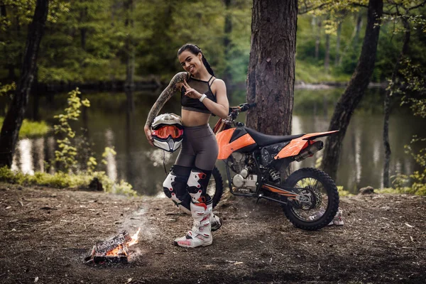 Charmante junge Rennfahrerin im Motocross-Outfit mit halbnacktem Oberkörper, Helm haltend und vor einer Kamera am Lagerfeuer im Wald posierend — Stockfoto