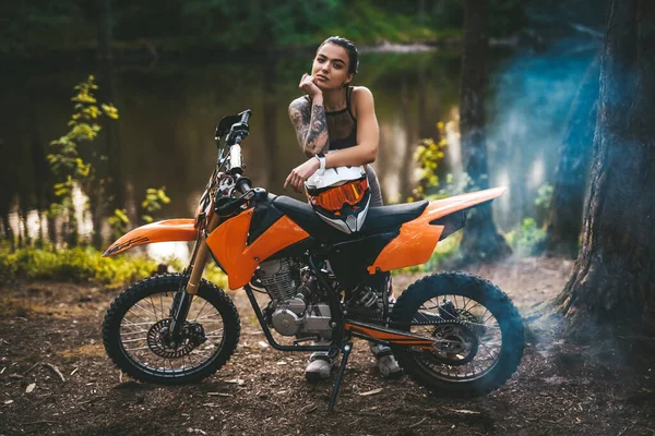 Urocza wytatuowana dziewczyna wyścigowa w motocrossowym stroju z półnagim tułowiem opierającym się na rowerze i patrzącym na kamerę w lesie — Zdjęcie stockowe