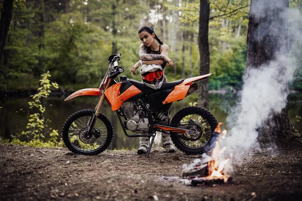 Affascinante giovane pilota donna vestita da motocross con busto semi nudo appoggiato alla sua moto e guardando su una macchina fotografica accanto al falò nel bosco — Foto Stock
