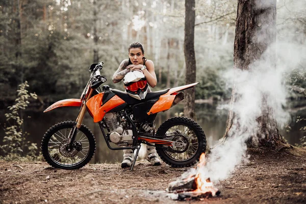 Charmante junge Rennfahrerin im Motocross-Outfit mit halbnacktem Oberkörper lehnt auf ihrem Fahrrad und blickt auf eine Kamera am Lagerfeuer im Wald — Stockfoto