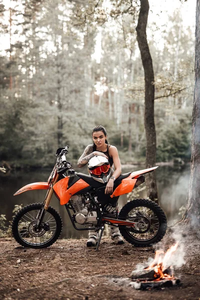 Urocza młoda zawodniczka ubrana w strój motocrossa z półnagim tułowiem opierającym się na rowerze i patrzącym na kamerę obok ogniska w lesie — Zdjęcie stockowe